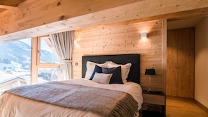 Et soveværelse med træloft, en stor seng med hvide og mørke puder, et sengebord med en lampe og et vindue med gardiner.