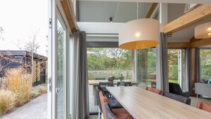 A modern dining area with a long wooden table, cushioned benches, and a large hanging light fixture. - Zonnedorp 21 Beach Villa Joya Renesse Luxury Beach Villa With Sauna (Renesse)