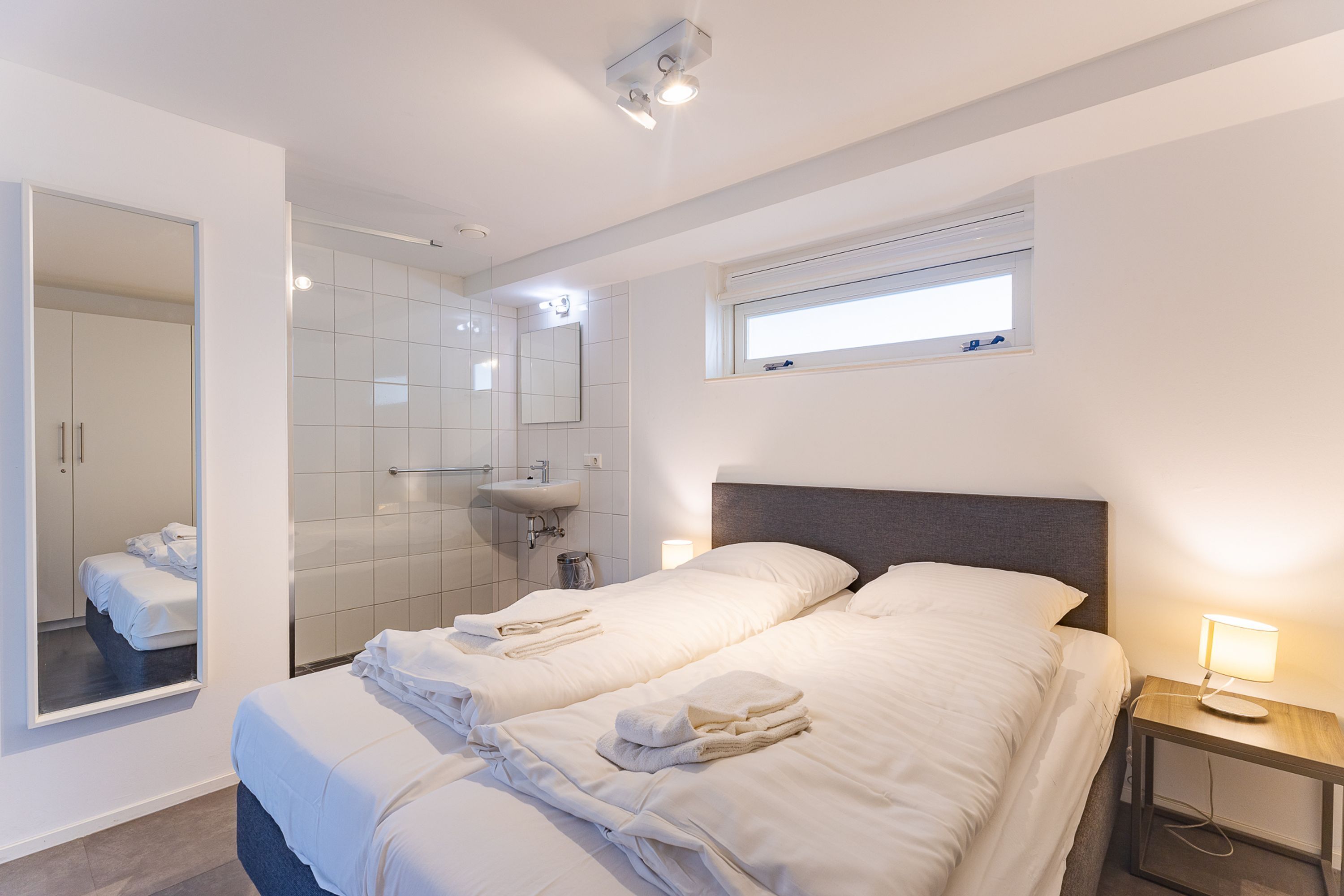 Een hotelkamer met twee bedden, een grote spiegel, een raam, een nachtkastje met lamp en een badkamer met wastafel en spiegel.