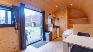 A wooden cabin interior with a dining table, chairs, a bed, a small kitchen area, and a sliding glass door leading to an outdoor patio. - Duinhoeve Duinlodges 4pers Huisdiervriendelijk (Burgh-Haamstede)