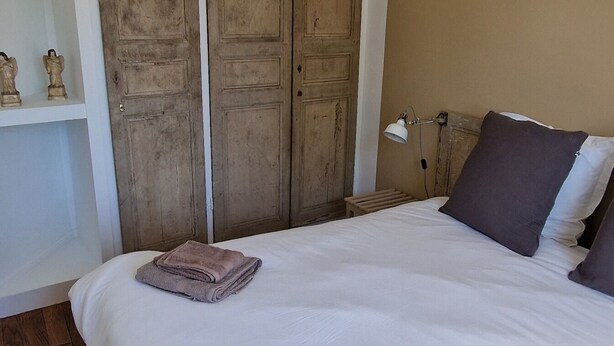 Un dormitorio con una cama grande, dos toallas dobladas, una lámpara colgante, un armario de madera y una mesita de noche con lámpara.