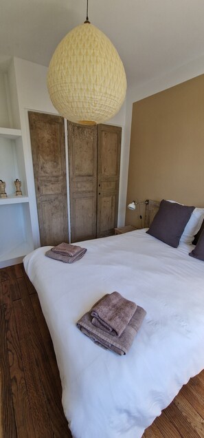 Une chambre avec un grand lit, deux serviettes pliées, un luminaire suspendu, une armoire en bois et une table de chevet avec une lampe.