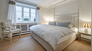 Et soveværelse med en stor seng, en stol, et natbord med en lampe og et vindue med gardiner.