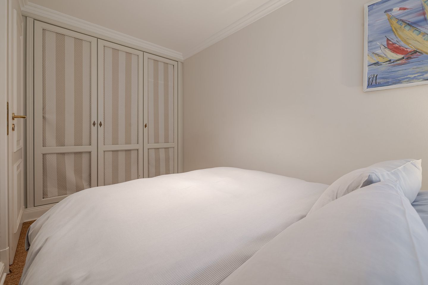 Quarto com um guarda-roupa branco grande, uma cama com roupa de cama branca e um quadro na parede.
