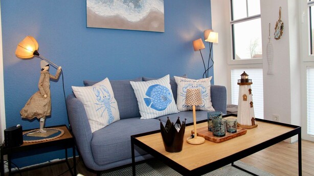 A room with a blue wall, a grey sofa, a wooden coffee table, a lamp, and a small lighthouse decoration. - Deichkrone - New Deichkrone vacation apartment with harbor view, balcony & (Krummhörn)