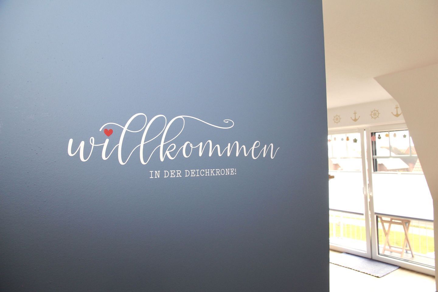 Un mur orné d’une pancarte d’accueil en allemand, une porte et une vue sur un balcon avec une chaise.