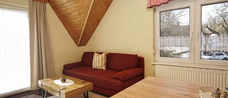 A cozy room with a wooden ceiling, a red sofa, a wooden table, and a window with a view of a snowy landscape.