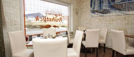 Un restaurant offrant une vue sur un port, avec des chaises blanches, des tables en bois et un grand tableau représentant un voilier.