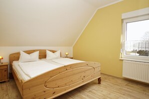 Un dormitorio con cama de madera, ropa de cama blanca, una mesita de noche con lámpara, una ventana con vistas y un radiador.