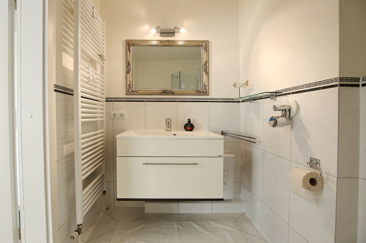 Een badkamer met een wit wastafel, een spiegel met een sierlijk lijstwerk, een handdoekhouder en een toiletrolhouder.