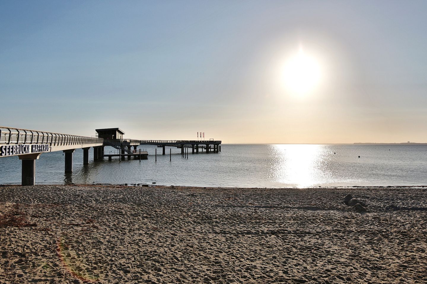 Een strand met een pier die zich uitstrekt over kalm water, een heldere lucht en een zon die laag aan de horizon staat.
