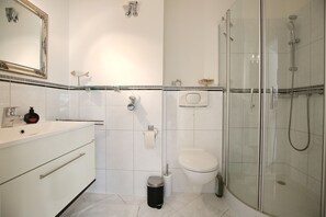 Un baño con ducha, inodoro y lavabo, que incluye un espejo, toallero y un cubo de basura.