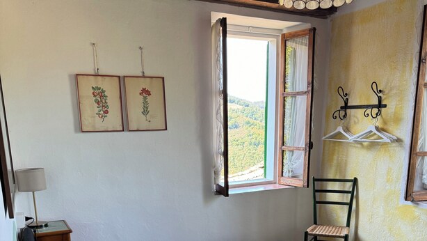 A bedroom with a bed, a chair, a table with a lamp, a window with a view, and wall-mounted hangers. - Casa del Falco - Borgo Spigone - Vetto d'Enza - Reggio Emilia (vetto)