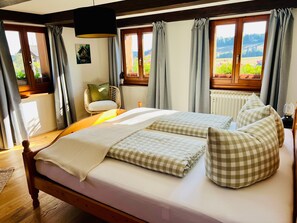 A double bed with checkered pillows, a wooden headboard, and a view of a garden through a window.