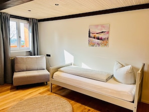 A bedroom with a bed, a chair, a window with curtains, a painting on the wall, and a wooden floor.