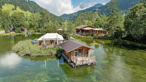 A floating wooden structure with a covered deck and a lifebuoy, surrounded by clear water and lush greenery.