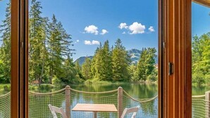 A wooden deck with a table and chairs overlooking a lake, surrounded by trees and mountains.