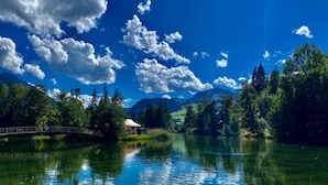 A serene lake with a wooden bridge, surrounded by lush greenery and mountains under a clear blue sky.