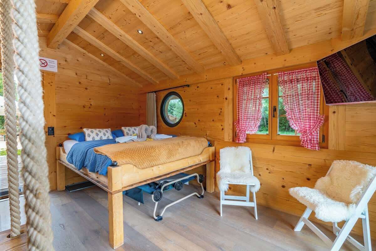 Un dormitorio de cabaña de madera con una cama, dos sillas, un ventanal con cortinas a cuadros y un ventanuilla circular sobre la cama.