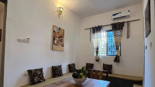 A dining area with a wooden table, chairs, and a bowl of fruit. There is a ceiling fan, a wall-mounted light fixture, and a window with curtains. - Cozy 3-bedroom Home 2 Minutes From Jambiani Beach (Jambiani)