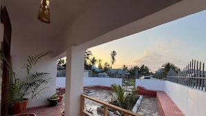 A balcony with potted plants, a railing, and a view of a residential area during sunset. - Cozy 3-bedroom Home 2 Minutes From Jambiani Beach (Jambiani)