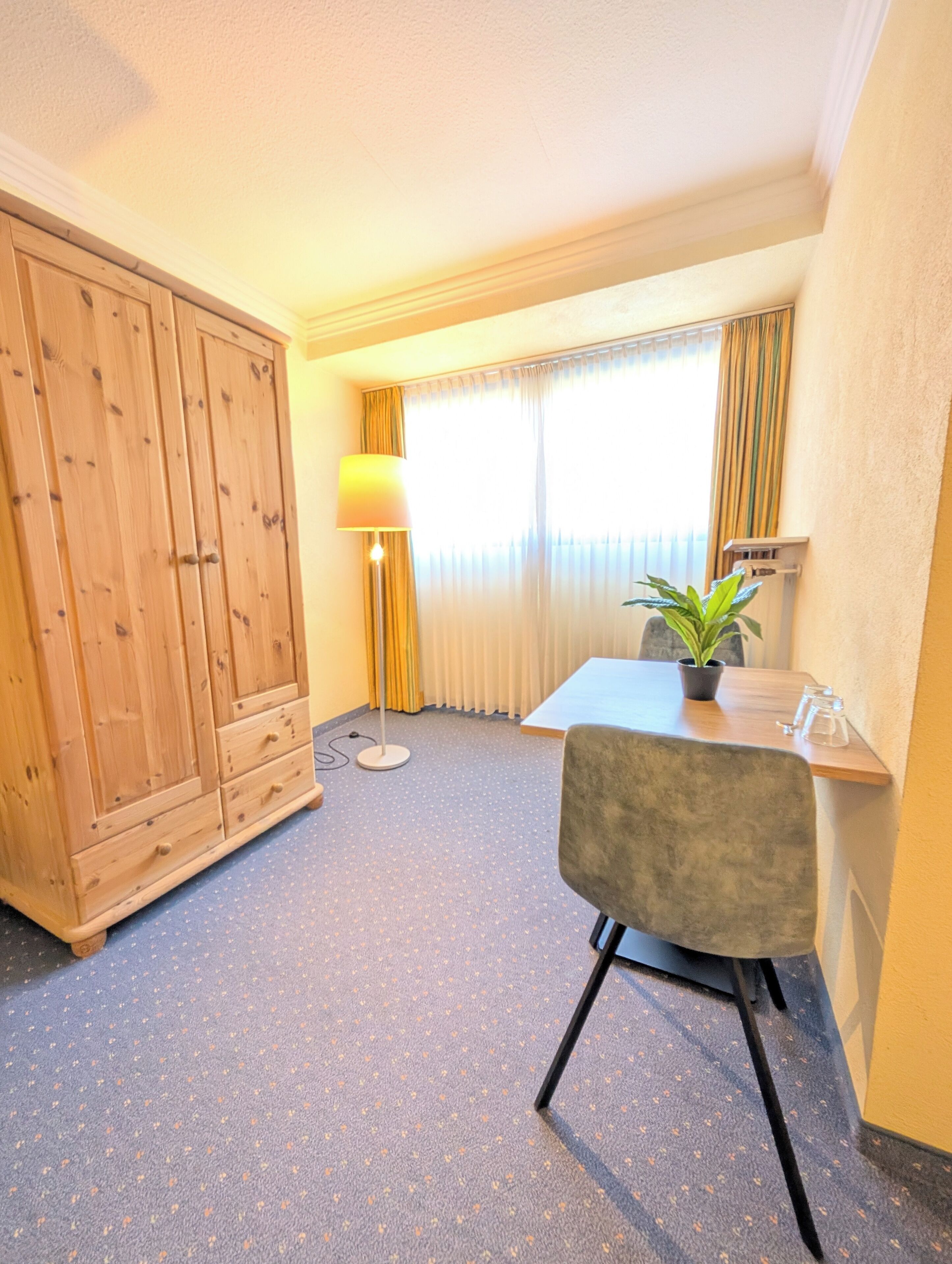 Une chambre d’hôtel avec une armoire en bois, un bureau avec une chaise, une plante en pot et une fenêtre avec des rideaux.
