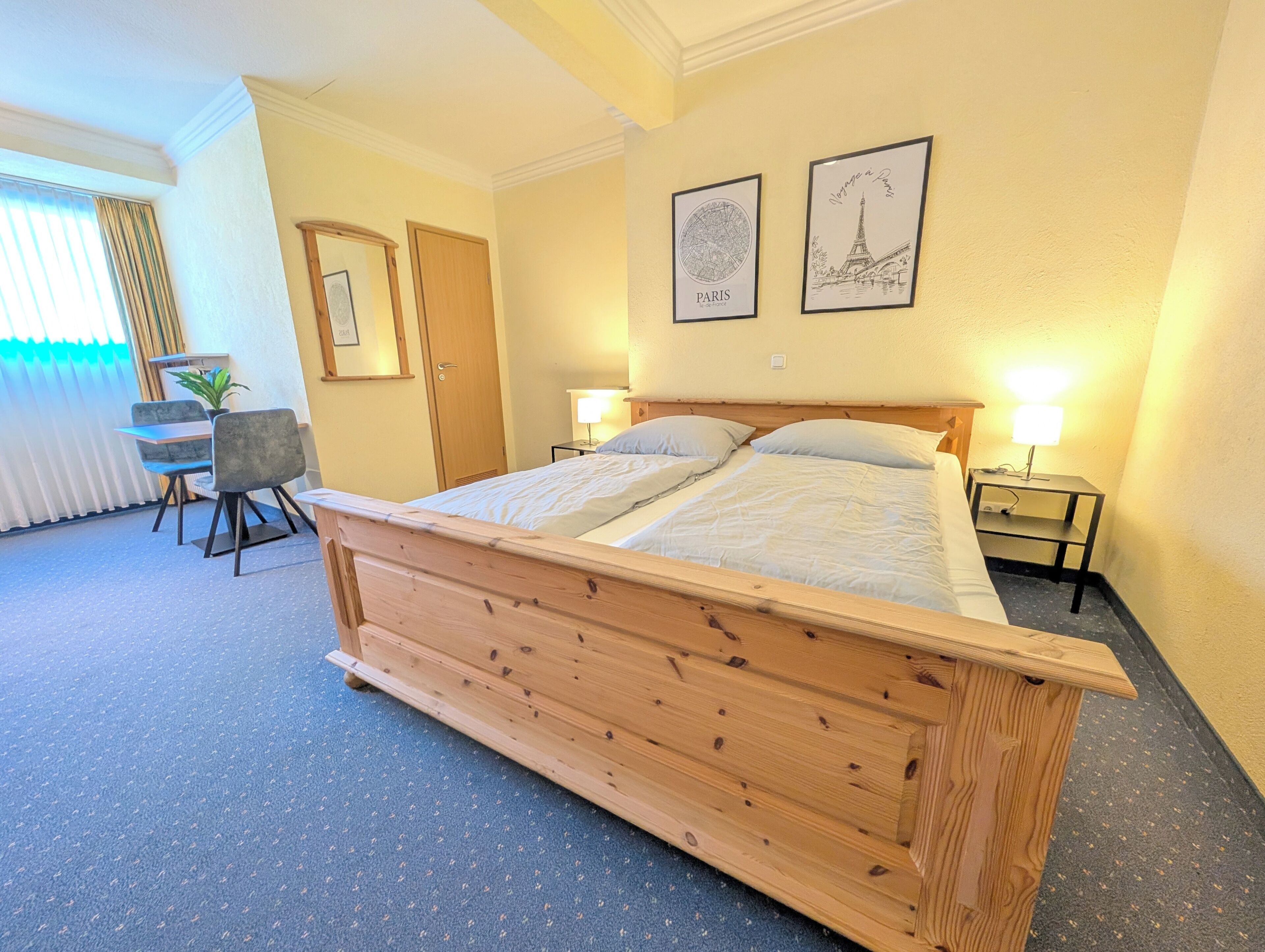 Un dormitorio con una cama de madera, un escritorio, dos lámparas de noche y dos cuadros en la pared.