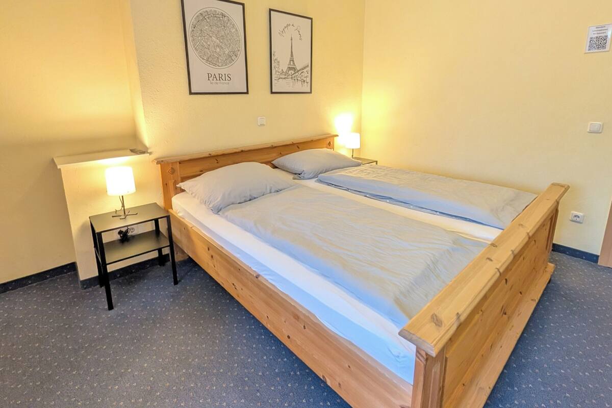 A bedroom with a wooden bed, two bedside lamps, a small table, and two framed pictures on the wall.