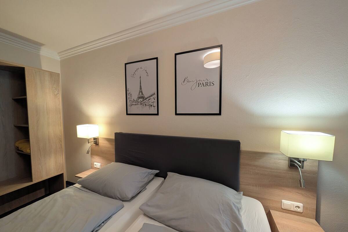 A hotel room with a bed, two bedside lamps, a nightstand, a wooden shelf, and two framed pictures on the wall.