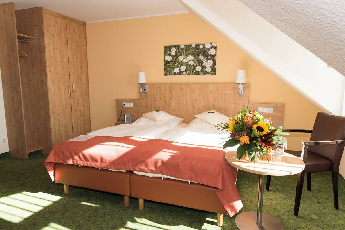 A hotel room with a bed, a desk with a chair, a wooden wardrobe, and a wall-mounted lamp.
