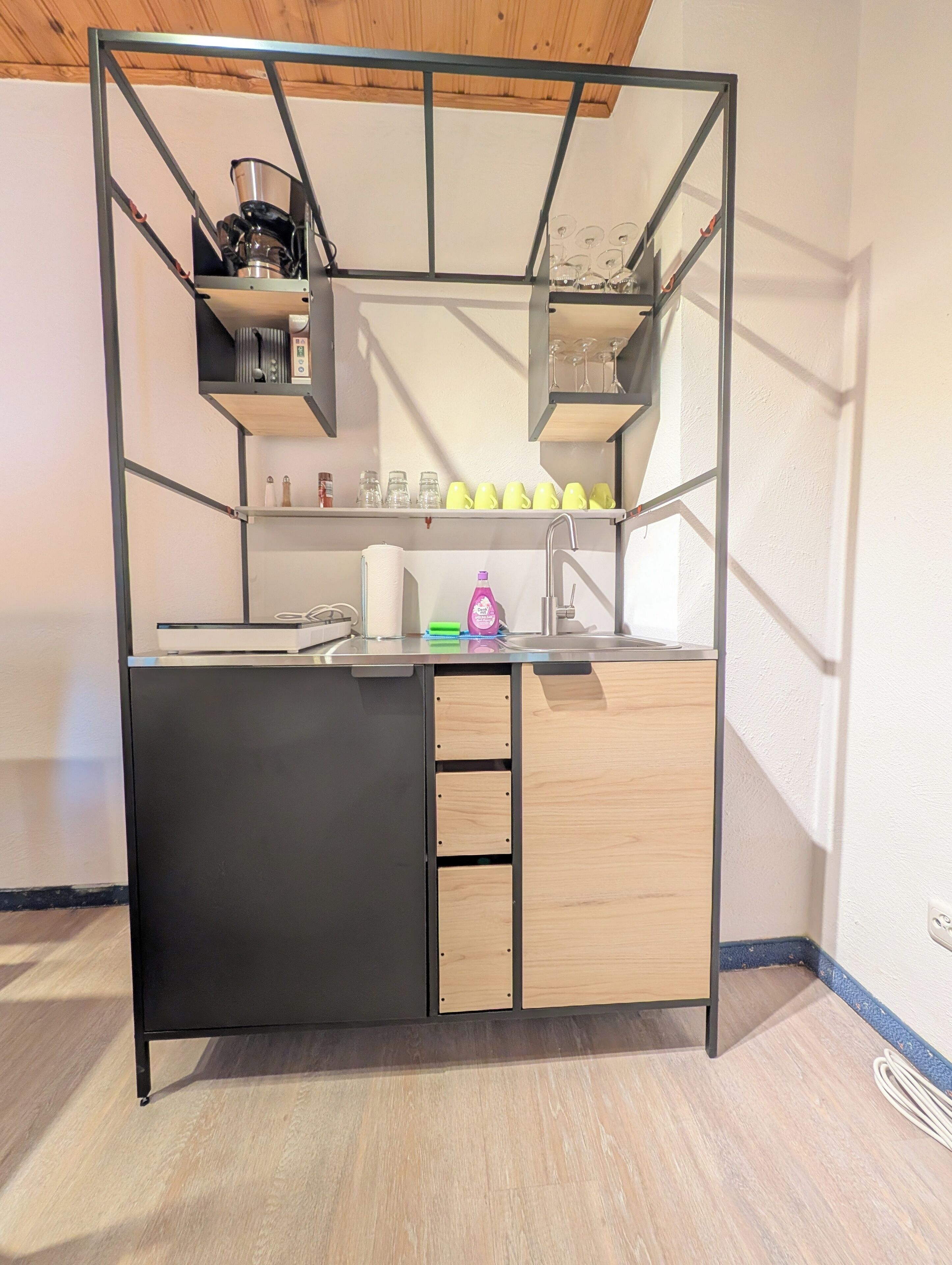A compact kitchenette with a black countertop, wooden cabinets, and a metal frame structure.