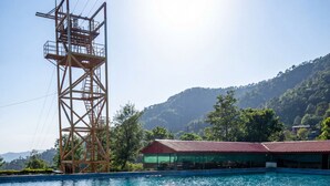 A swimming pool with a wooden structure in the background, surrounded by a building with a red roof. - Mhara Himachal (Bir)