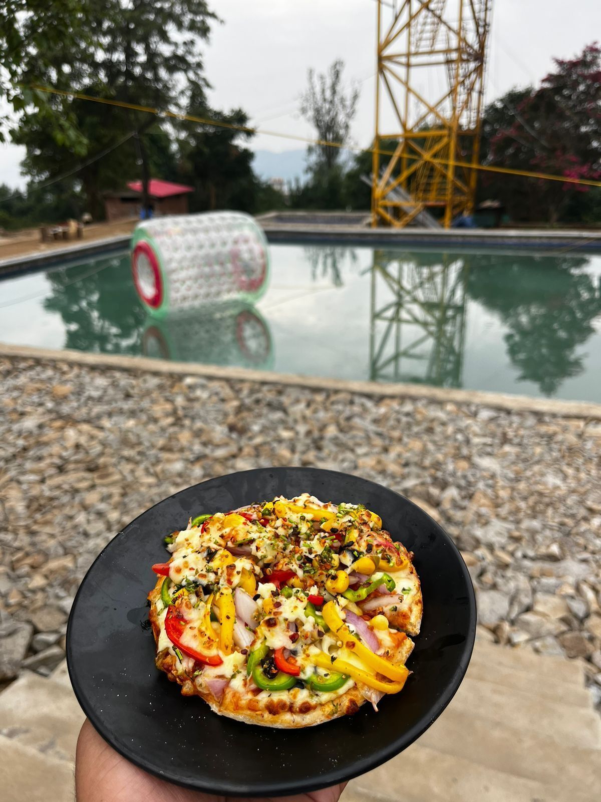A pizza on a black plate held in front of a pool with a water slide and a tower structure in the background.