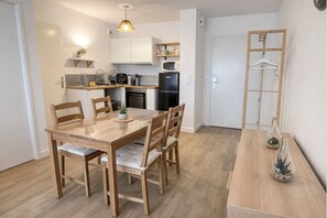 A compact kitchen with white cabinets, a microwave, and a refrigerator. There is a wooden dining table with chairs and a hanging light fixture above it. - Cosy apartment with balcony 15 minutes from Lille (Ronchin)