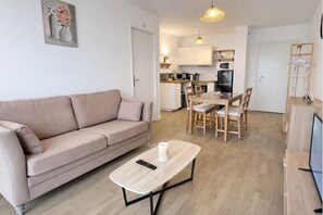 A modern living room with a beige sofa, a wooden coffee table, and a dining area with a wooden table and chairs. A kitchen is visible in the background with white cabinets and a microwave. - Cosy apartment with balcony 15 minutes from Lille (Ronchin)