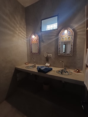 Un baño con dos lavamanos, cada uno con un espejo decorativo y una toalla. Hay un pequeño estante con una caja de pañuelos y un jabonero.
