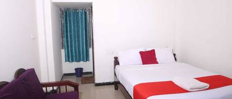 A hotel room with a bed, a chair, a small table, a window with curtains, and an air conditioning unit.
