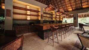 A bar area with wooden shelves and stools, a thatched roof, and a view of greenery outside. - Naio Hotel (Dibulla)