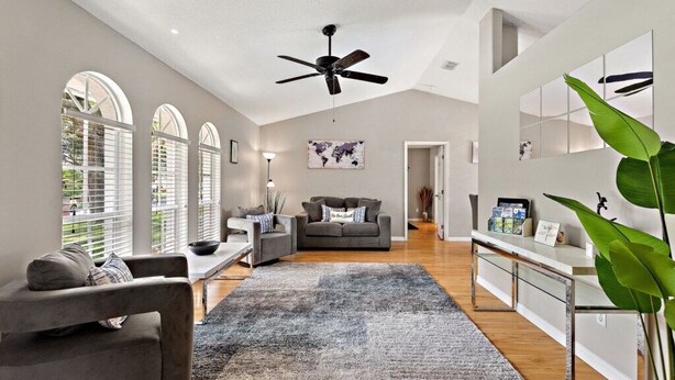 Ein modernes Wohnzimmer mit grauem Sofa, einem Couchtisch und einem großen Teppich. Es gibt einen Deckenventilator, eine Weltkarte an der Wand und eine Pflanze in der Ecke.