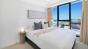 A hotel room with a bed, a ceiling fan, a nightstand, a lamp, and a large window with a view of the beach and ocean. - Absolute Beachfront on Albatross - SeaView 3 bed (Mermaid Beach)