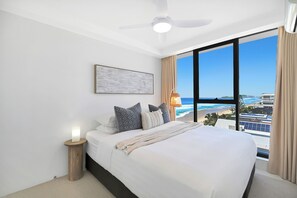 A hotel room with a bed, a ceiling fan, a nightstand, a lamp, and a large window with a view of the beach and ocean. - Absolute Beachfront on Albatross - SeaView 3 bed (Mermaid Beach)
