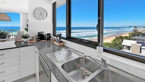 A modern kitchen with a large window overlooking a beach, a clock on the wall, and a view of the ocean. - Absolute Beachfront on Albatross - SeaView 3 bed (Mermaid Beach)