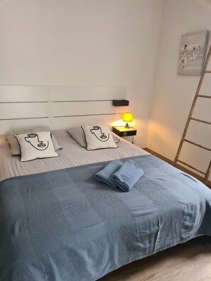 A neatly made bed with a blue blanket and two pillows, a bedside lamp, and a ladder against the wall. - House downtown (Dol-de-Bretagne)