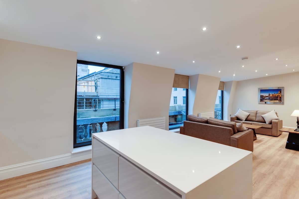 A modern living room with a large window, a sofa set, and a kitchen area.