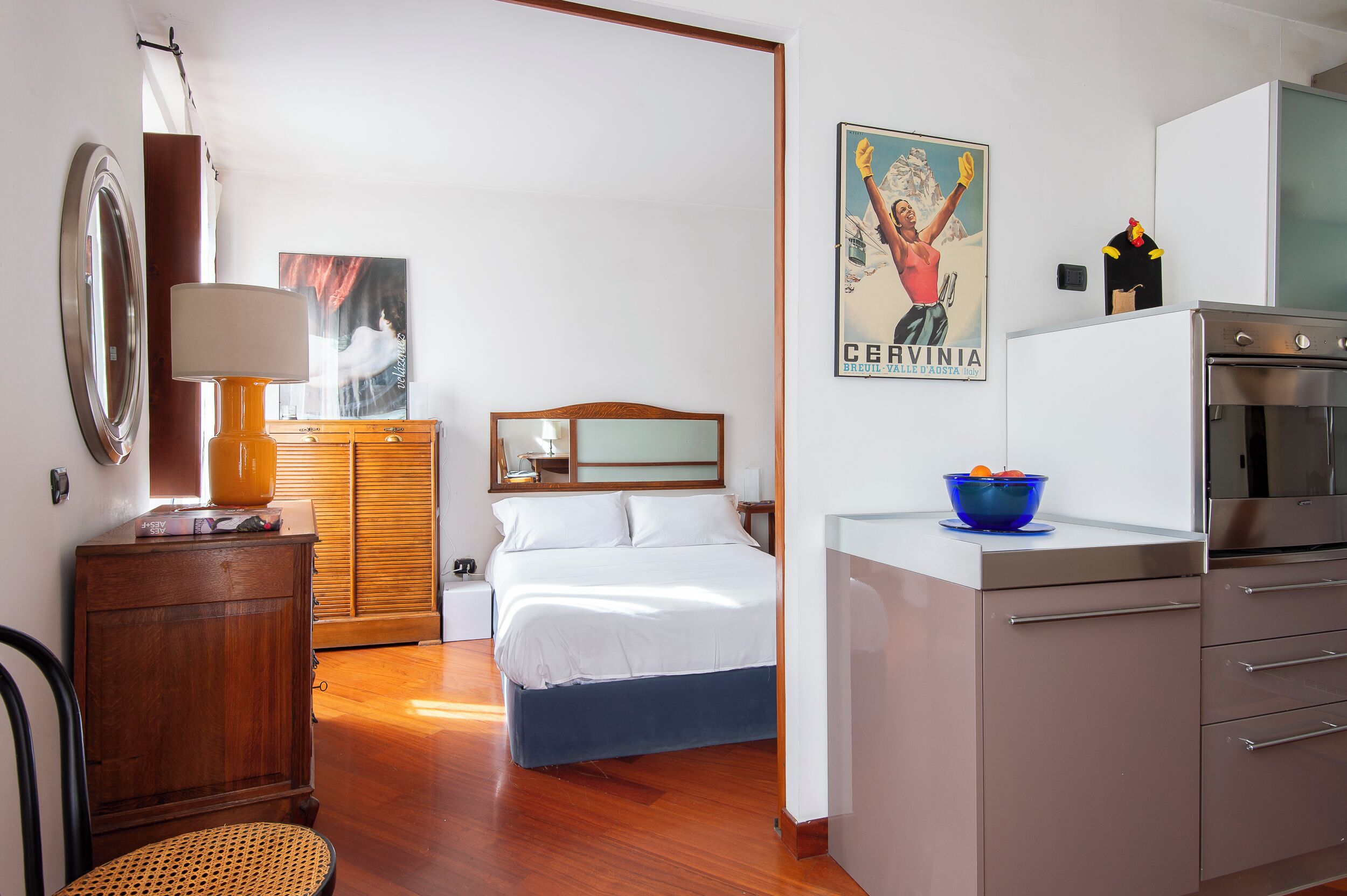 Ein modernes Hotelzimmer mit Bett, einem Holzschrank, einem Schreibtisch mit Lampe, einer Küchenzeile mit Kühlschrank und Herd sowie einem Poster an der Wand.