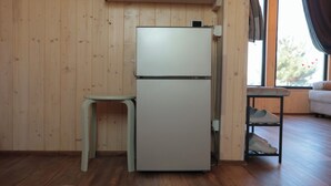 Ein gemütlicher Raum mit Holzboden, einem weißen Kühlschrank, einem kleinen Hocker und einem Fenster mit Blick auf Bäume.