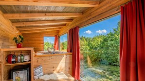Eine Hütte mit Holzvertäfelung, einer kleinen Küchenzeile, einem Fenster mit roten Vorhängen und Blick auf ein bewaldetes Gebiet.