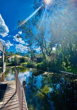 Una terraza de madera con vista a una masa de agua tranquila, con reflejos de árboles y un cielo azul despejado.