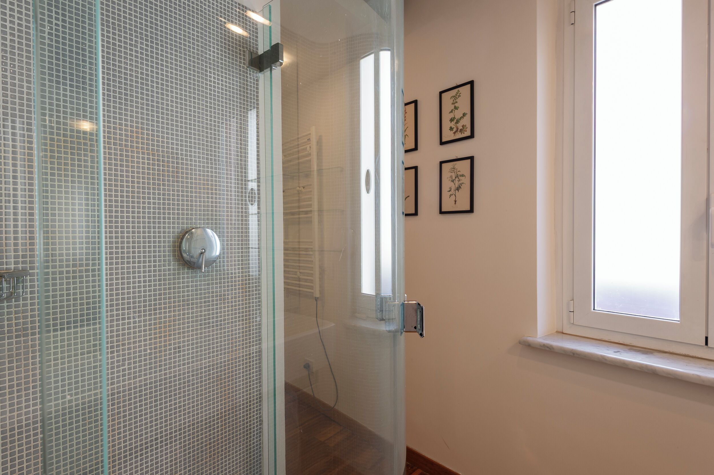 A bathroom with a glass shower enclosure, a white tiled wall, and a window with blinds.