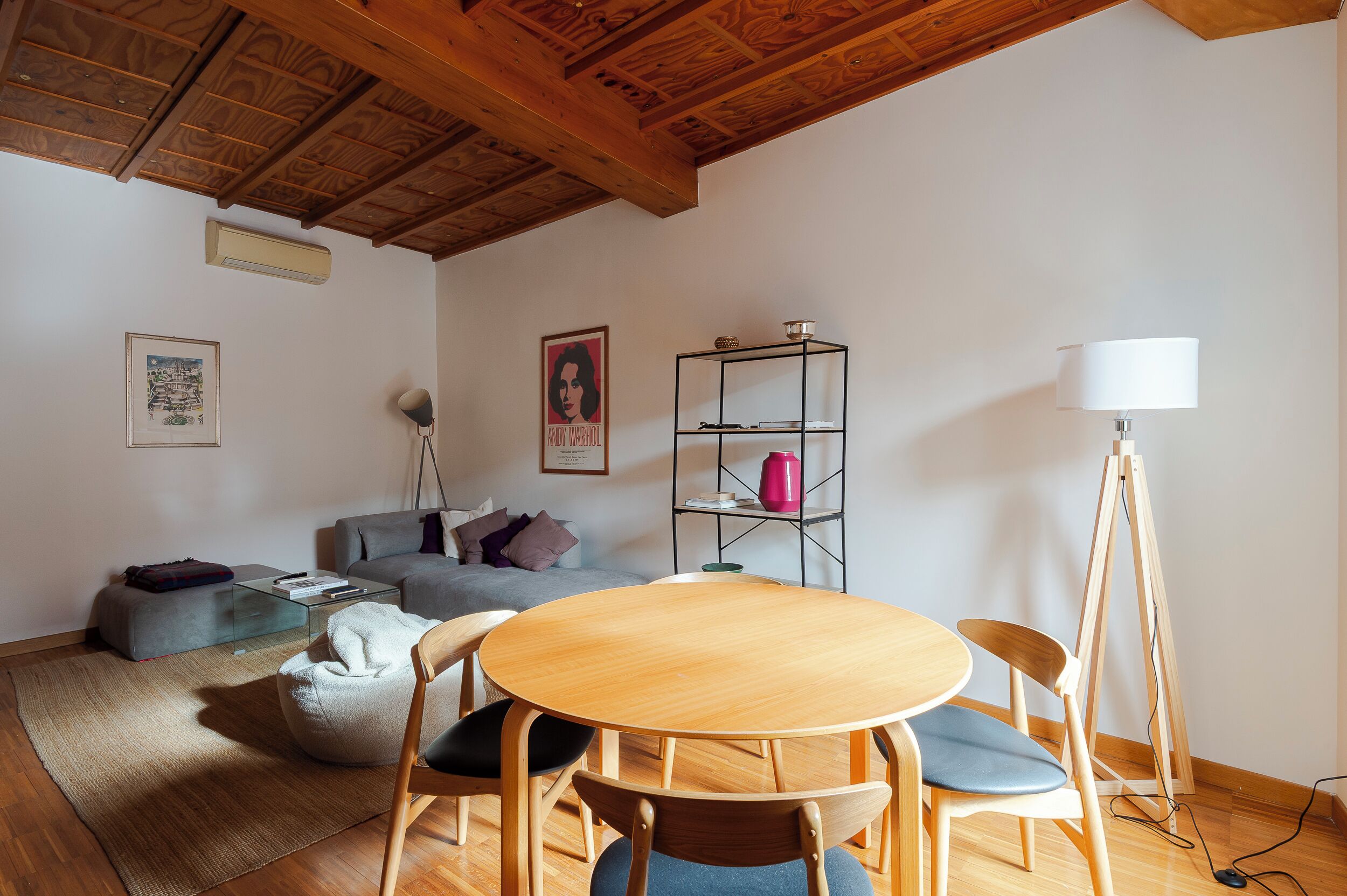 Un salón con techo de madera, una mesa redonda de madera con sillas, un sofá gris, una mesa de centro de vidrio, una estantería de metal y una lámpara de pie.